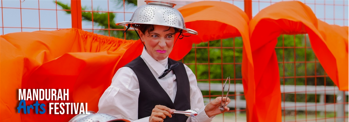 A woman wearing a colander on her head and holding tongs, standings within a bright red cage, with a comical expression on her face